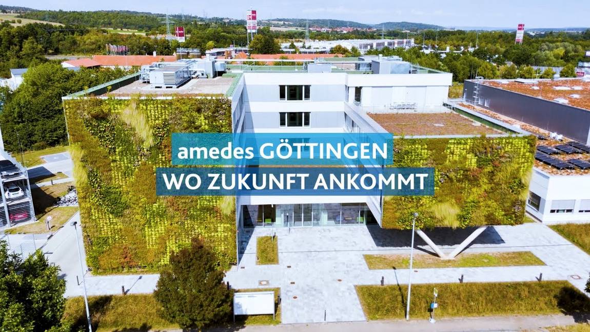 Ein modernes weißes Gebäude mit grünen Wänden und Pflanzen, umgeben von Bäumen und Grünpflanzen. Der überlagerte Text lautet: amedes GÖTTINGEN WO ZUKUNFT ANKOMMT.